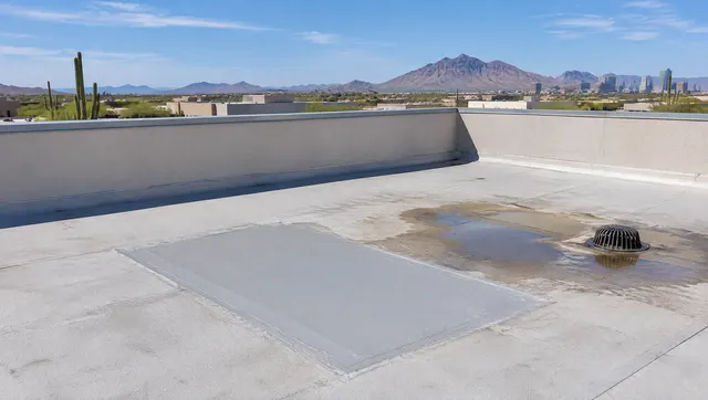 Wide flat commercial roof with parapet walls and a roof drain, a freshly repaired patched section visible near a ponding stain, Phoenix desert sky